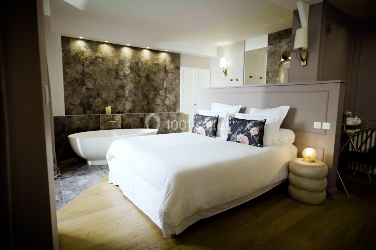 Chambre élégante avec lit double, baignoire blanche sur fond de mur texturé et éclairage tamisé.