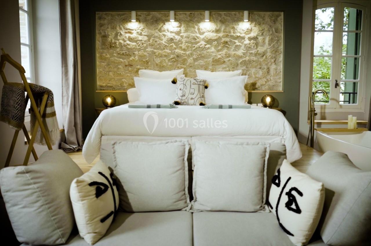 Chambre élégante avec lit double, canapé clair, mur en pierre éclairé et baignoire près d'une fenêtre lumineuse.