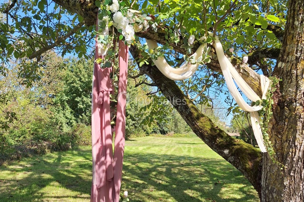 Tissu rose et rubans beige et bleu suspendus à une branche d'arbre dans un jardin ensoleillé.