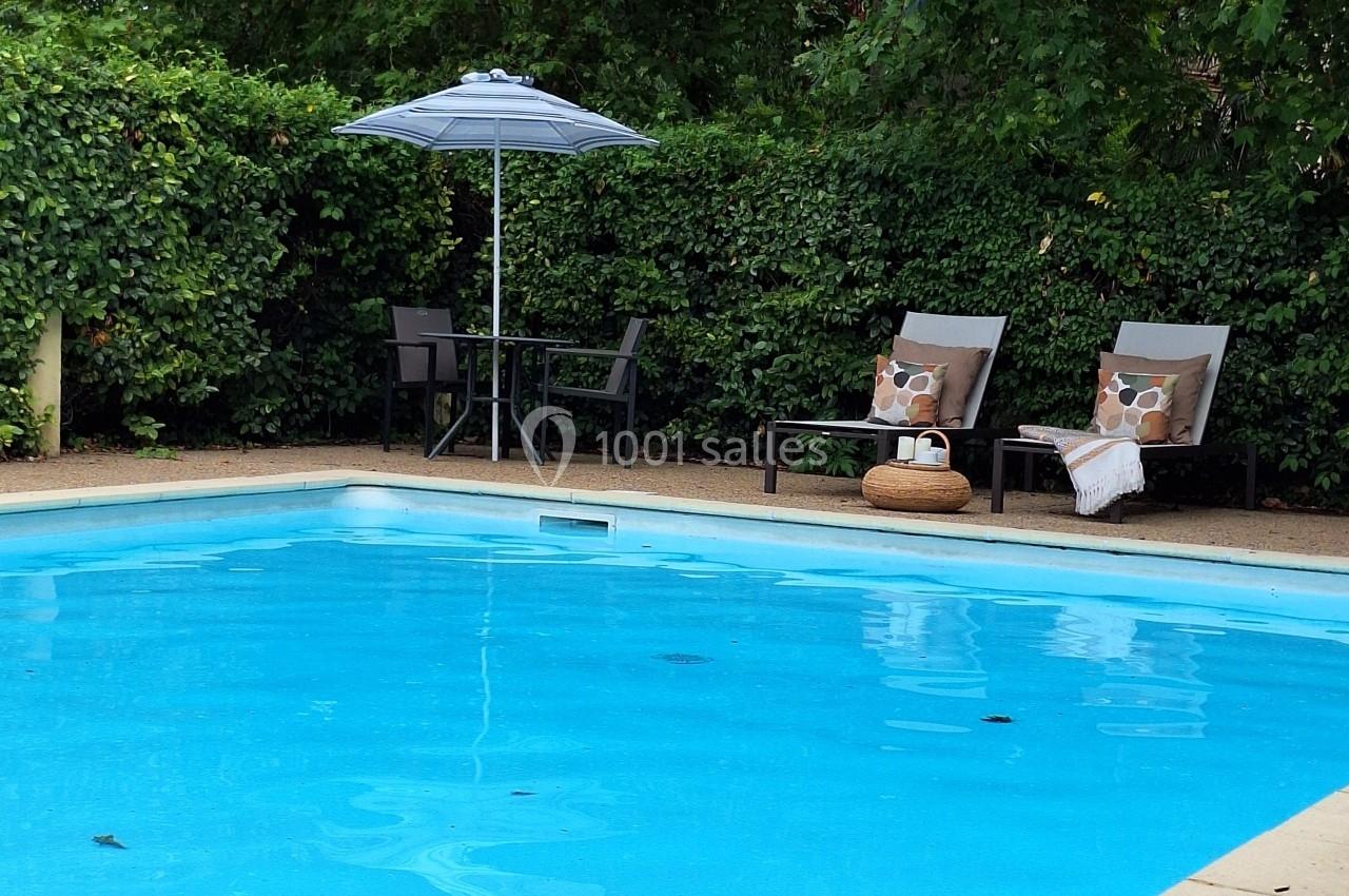 Piscine extérieure entourée de verdure avec deux chaises longues, un parasol et une table en arrière-plan.
