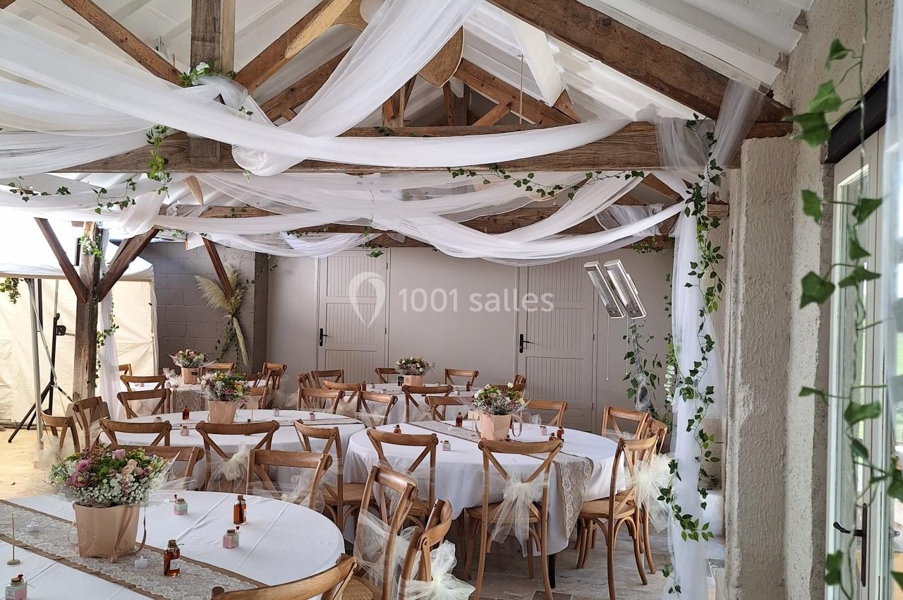 Salle de réception décorée avec des guirlandes blanches, des tables rondes ornées de fleurs et des chaises en bois.