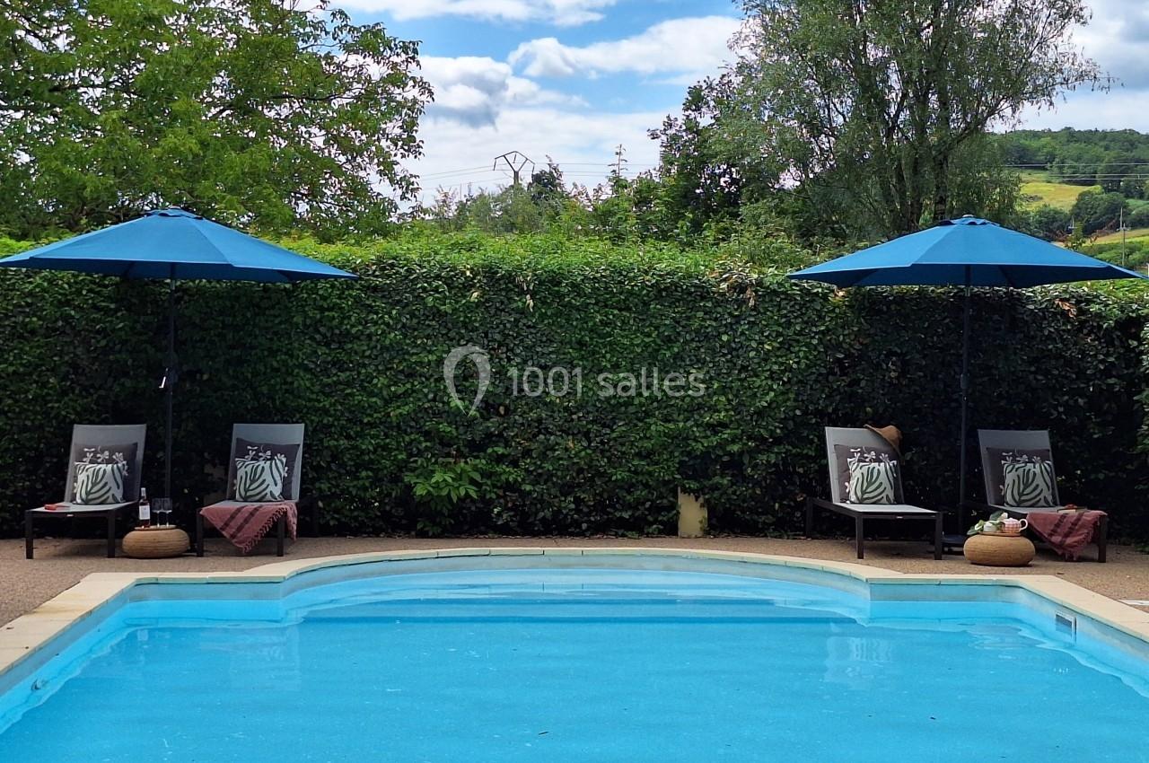 Piscine extérieure entourée de verdure, avec deux chaises longues et parasols bleus sur les côtés.