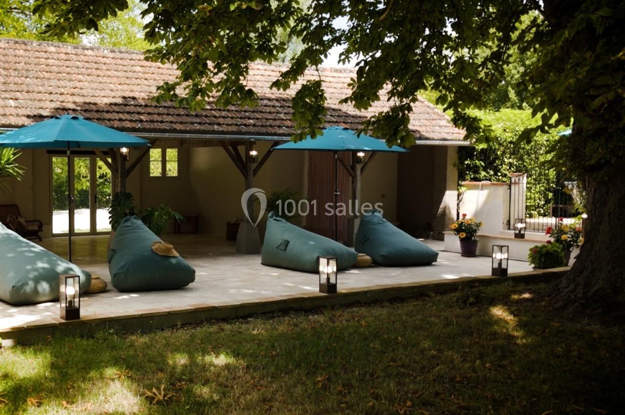 Terrasse ombragée avec des poufs verts, parasols bleus et éclairage, entourée de verdure et d'un bâtiment en arrière-plan.