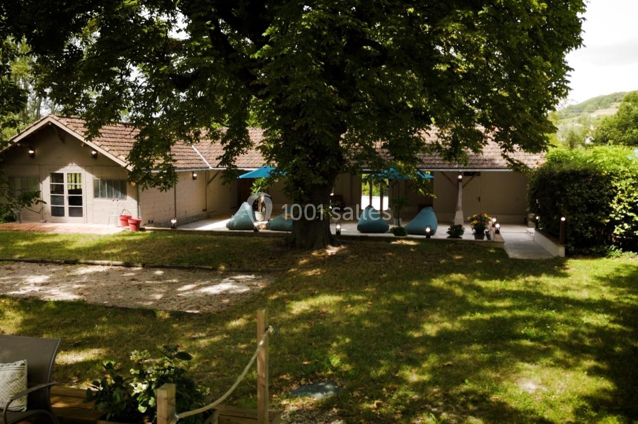 Maison de plain-pied entourée de verdure, avec une terrasse ombragée et des fauteuils sous un grand arbre.