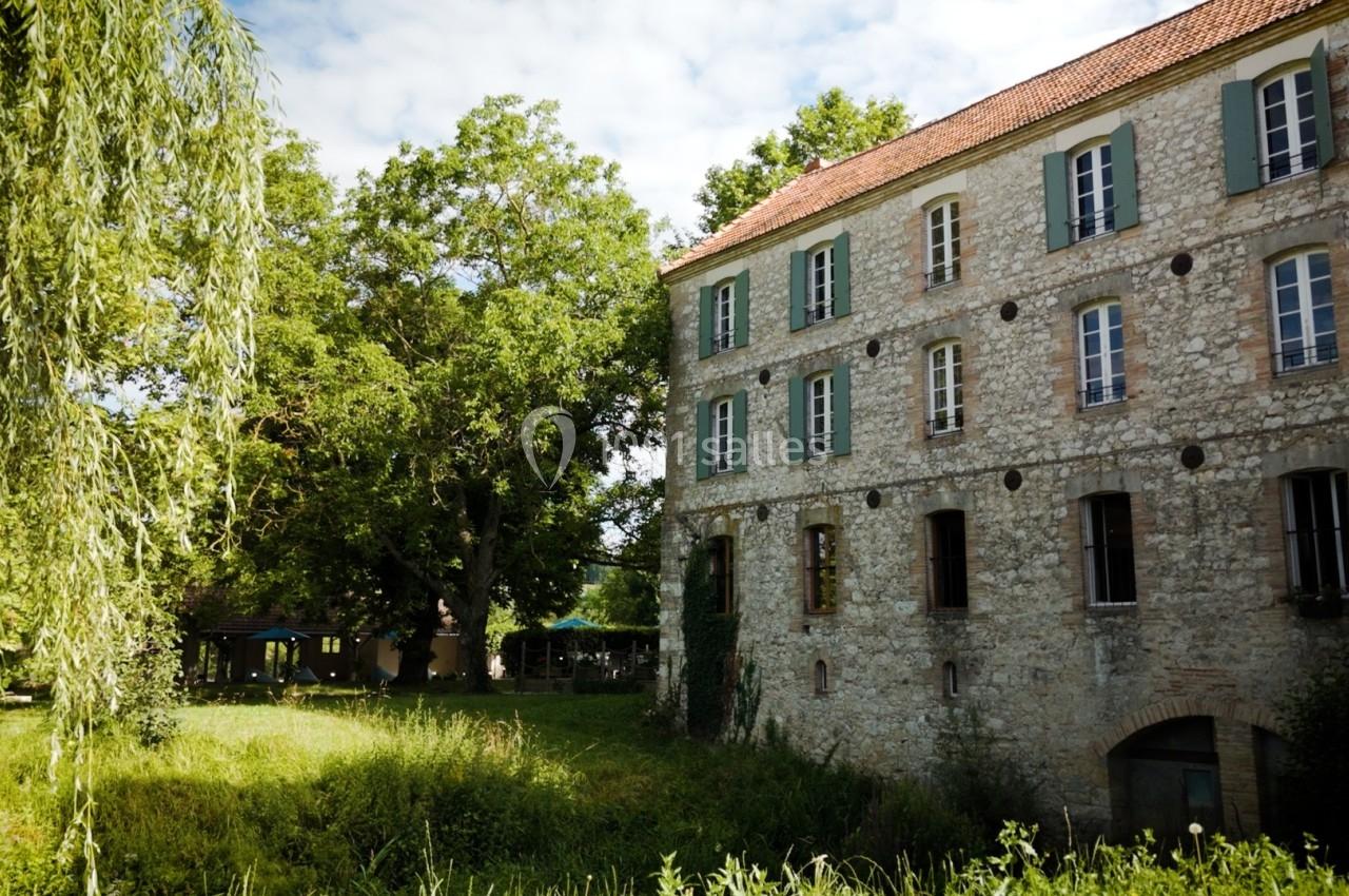 Bâtiment en pierre avec volets verts entouré d'arbres et d'herbe sous un ciel partiellement nuageux.