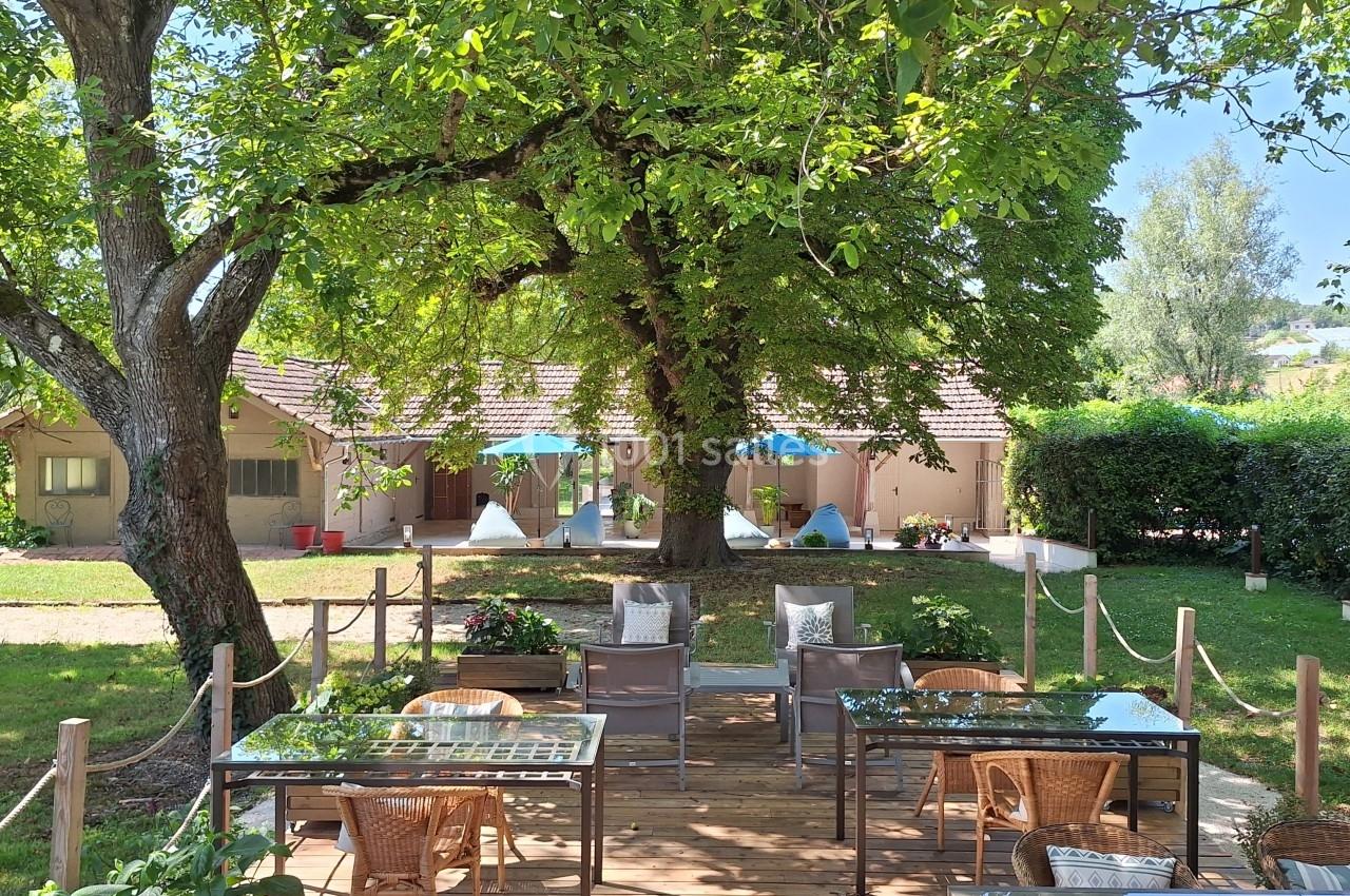 Terrasse en bois avec tables et chaises sous un grand arbre, entourée de verdure et de bâtiments clairs en arrière-plan.