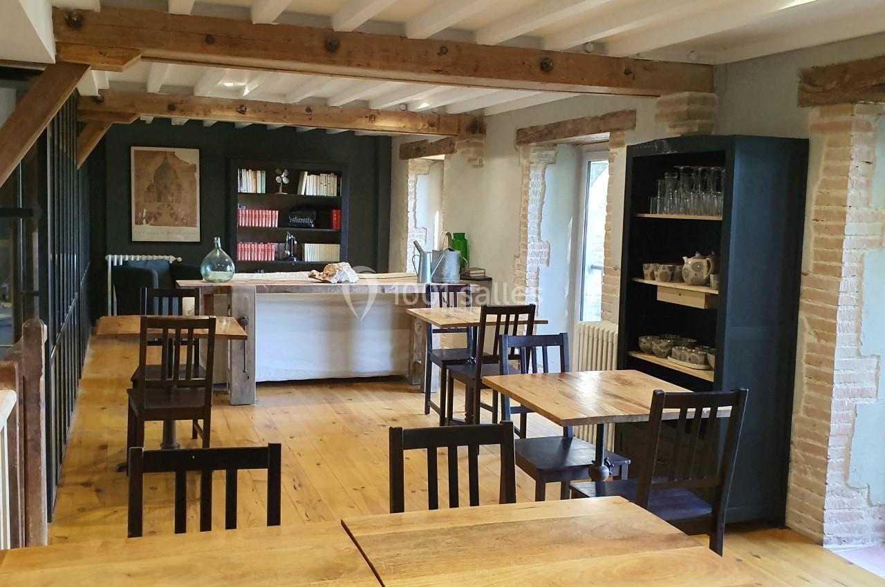 Salle lumineuse avec tables en bois, chaises sombres, étagères remplies et poutres apparentes au plafond.