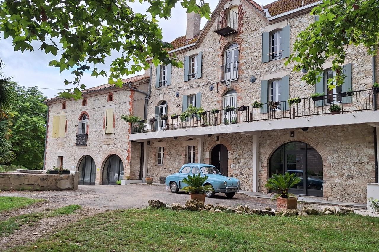 Façade d'une maison en pierre avec volets colorés, balcon fleuri, et une voiture ancienne stationnée devant.