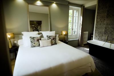 Miniature SUITE EUGENIE CHAMBRE 1 Chambre lumineuse avec lit double, fauteuil en bois, baignoire blanche et grande fenêtre donnant sur l'extérieur.