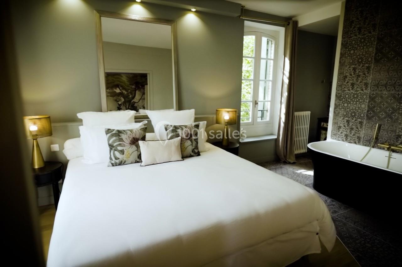 Chambre lumineuse avec lit double, baignoire indépendante, murs gris et grande fenêtre donnant sur l'extérieur.