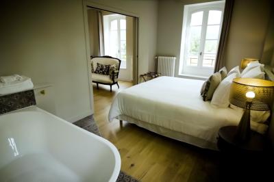 Miniature SUITE EUGENIE CHAMBRE 1 VERS SALON Chambre lumineuse avec lit double, fauteuil en bois, baignoire blanche et grande fenêtre donnant sur l'extérieur.