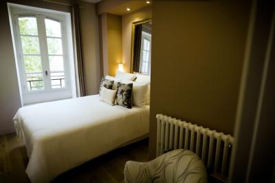 Miniature SUITE EUGENIE CHAMBRE 2 Chambre lumineuse avec lit double, fauteuil en bois, baignoire blanche et grande fenêtre donnant sur l'extérieur.