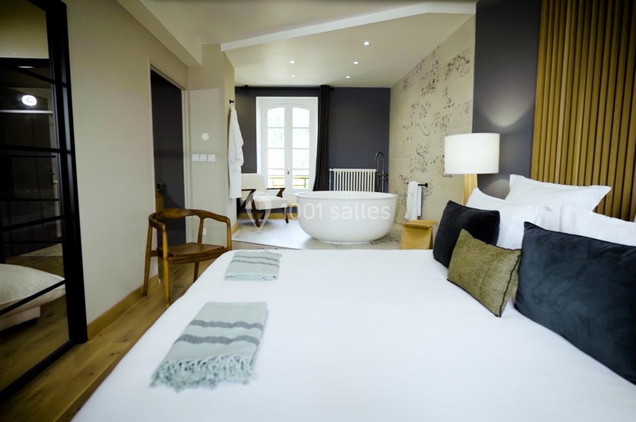 Chambre lumineuse avec lit double, fauteuil en bois, baignoire blanche et grande fenêtre donnant sur l'extérieur.