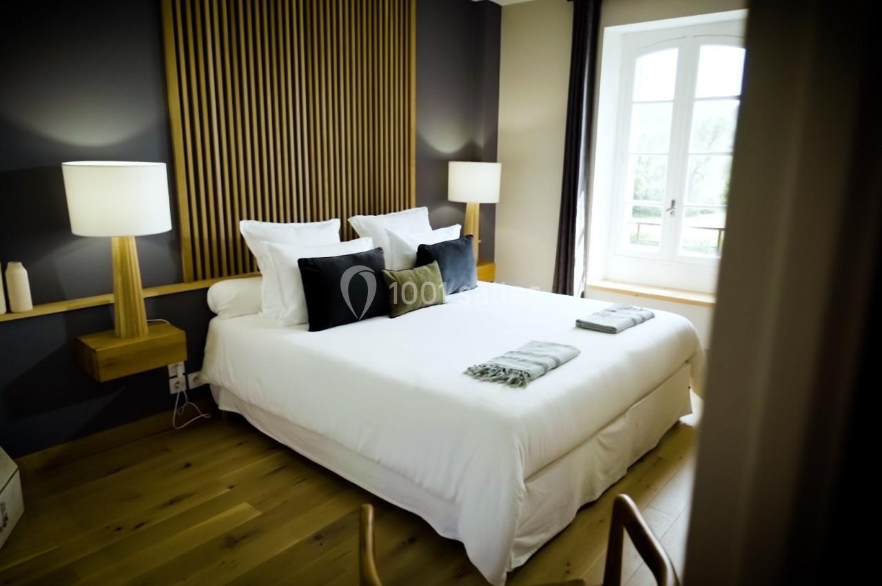 Chambre lumineuse avec lit double, linge blanc, coussins décoratifs, lampes modernes et grande fenêtre.