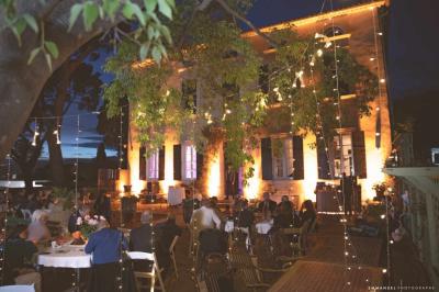 Façade d'une maison provençale jaune à volets verts, éclairée par le soleil couchant, avec une cour pavée et des plantes.