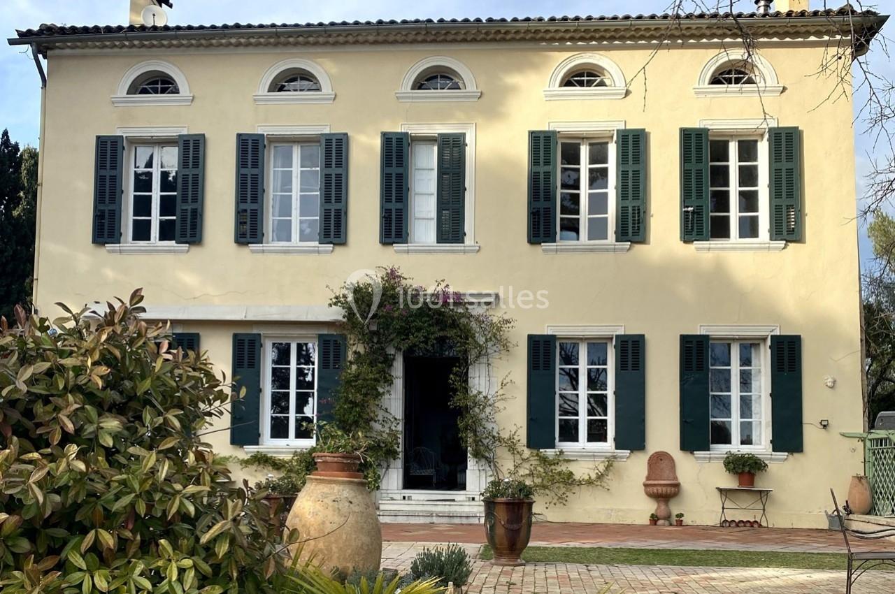 Façade d'une maison ancienne à deux étages, avec volets verts, entourée de végétation et d'éléments décoratifs.