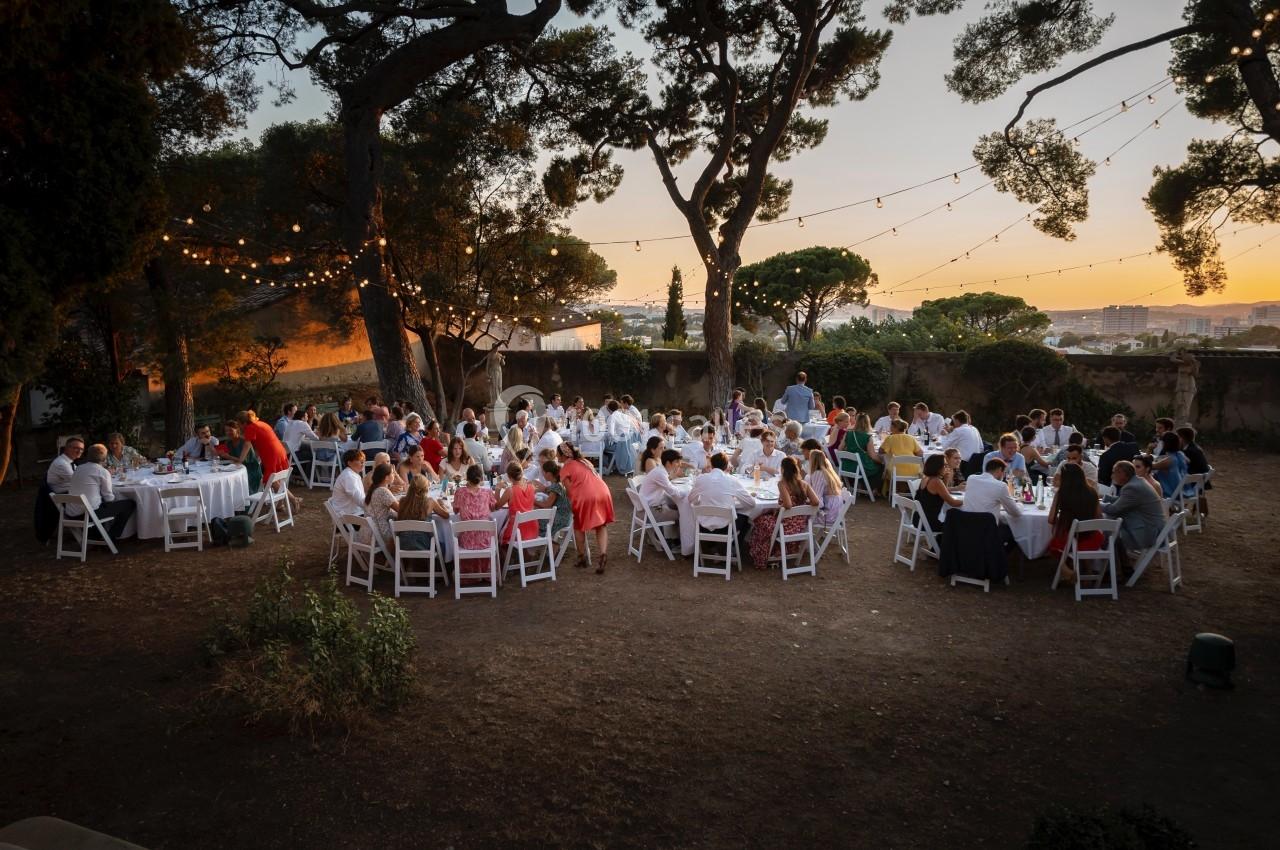 Groupe de personnes assises à des tables rondes en extérieur, sous des guirlandes lumineuses, au coucher du soleil.