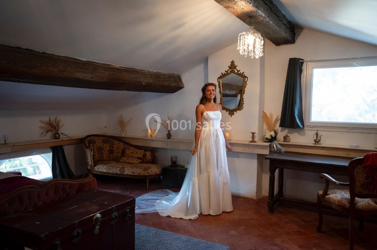 Une femme en robe de mariée se tient dans une chambre mansardée décorée de meubles anciens et d'éléments rustiques.