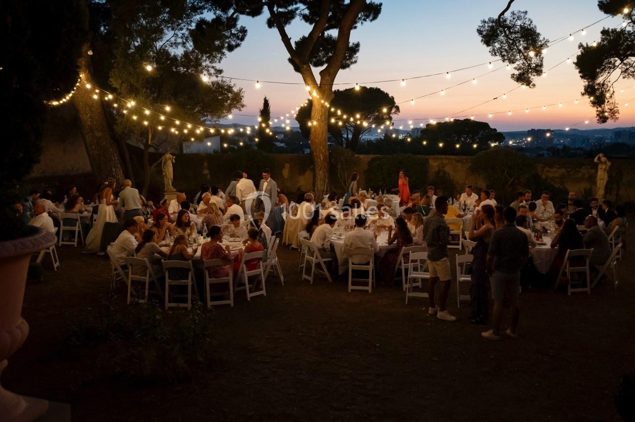 Dîner en plein air au crépuscule avec des invités assis à des tables décorées, éclairées par des guirlandes lumineuses.
