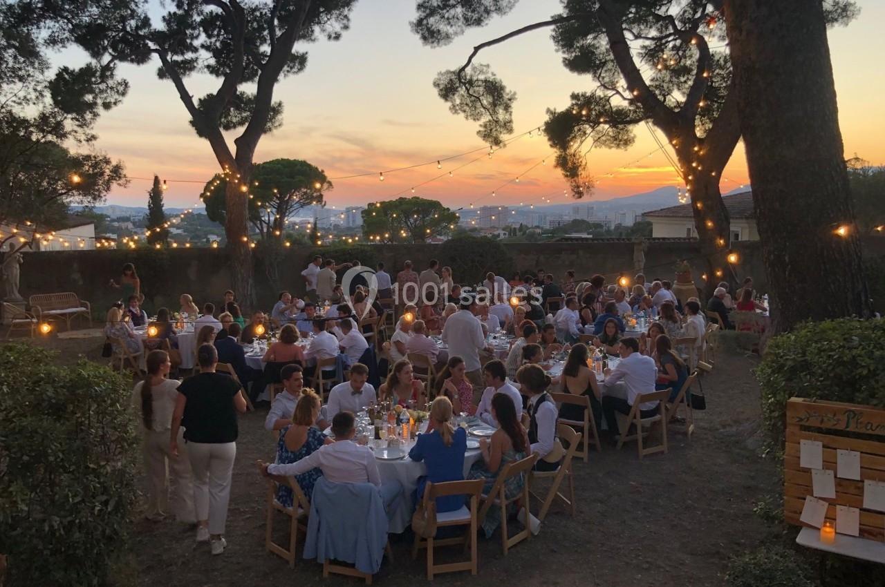Personnes dînant en plein air au coucher du soleil, entourées de guirlandes lumineuses et de grands arbres.