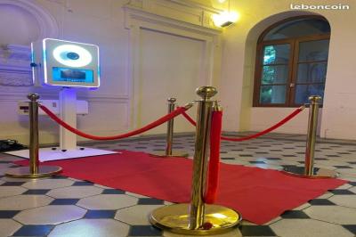 Borne photo interactive éclairée entourée de poteaux dorés et cordons rouges sur un tapis rouge dans une salle lumineuse.
