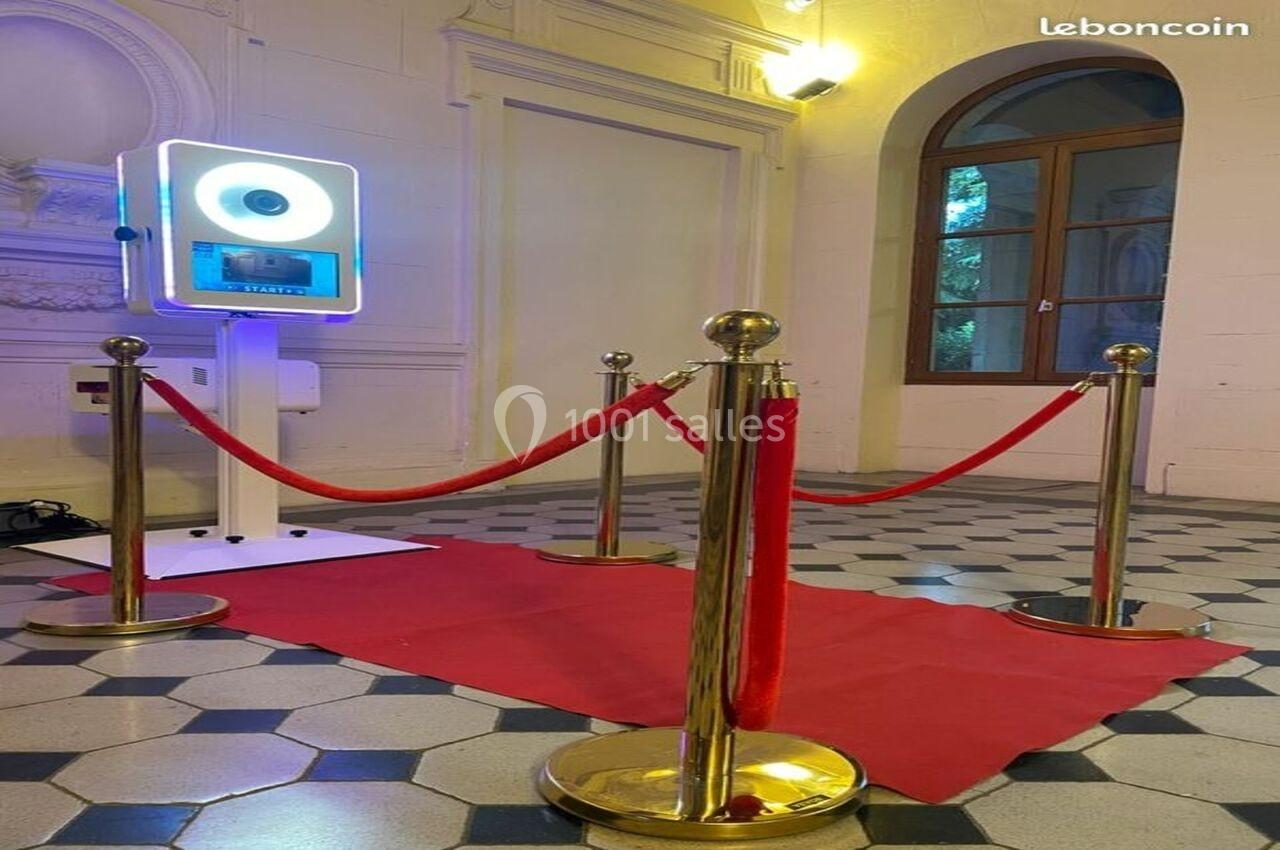 Borne photo interactive éclairée entourée de poteaux dorés et cordons rouges sur un tapis rouge dans une salle lumineuse.