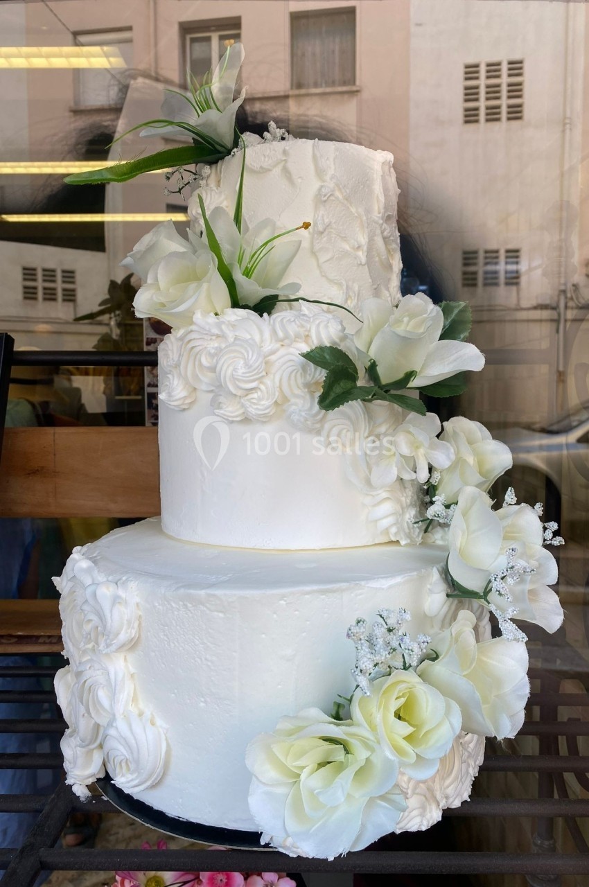 Gâteau de mariage à trois étages décoré de fleurs blanches et de motifs en crème, exposé en vitrine.