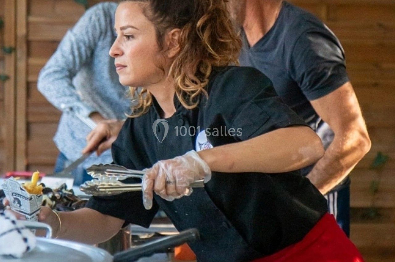 Une femme en tenue de cuisine sert un plat avec des pinces lors d'une activité en extérieur.