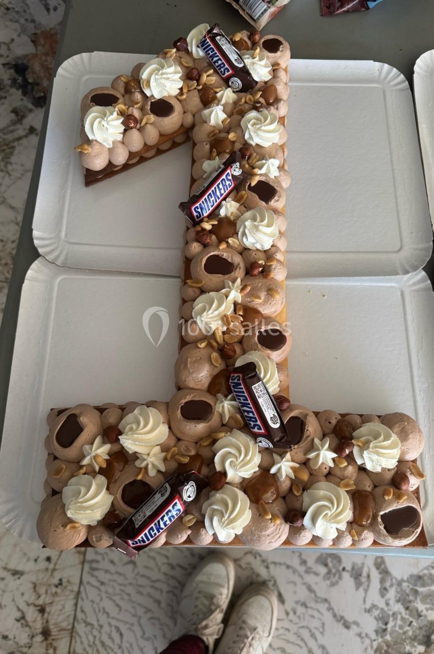 Gâteau en forme de chiffre 1 décoré de crème, cacahuètes, chocolat et mini barres Snickers, posé sur des assiettes blanches.