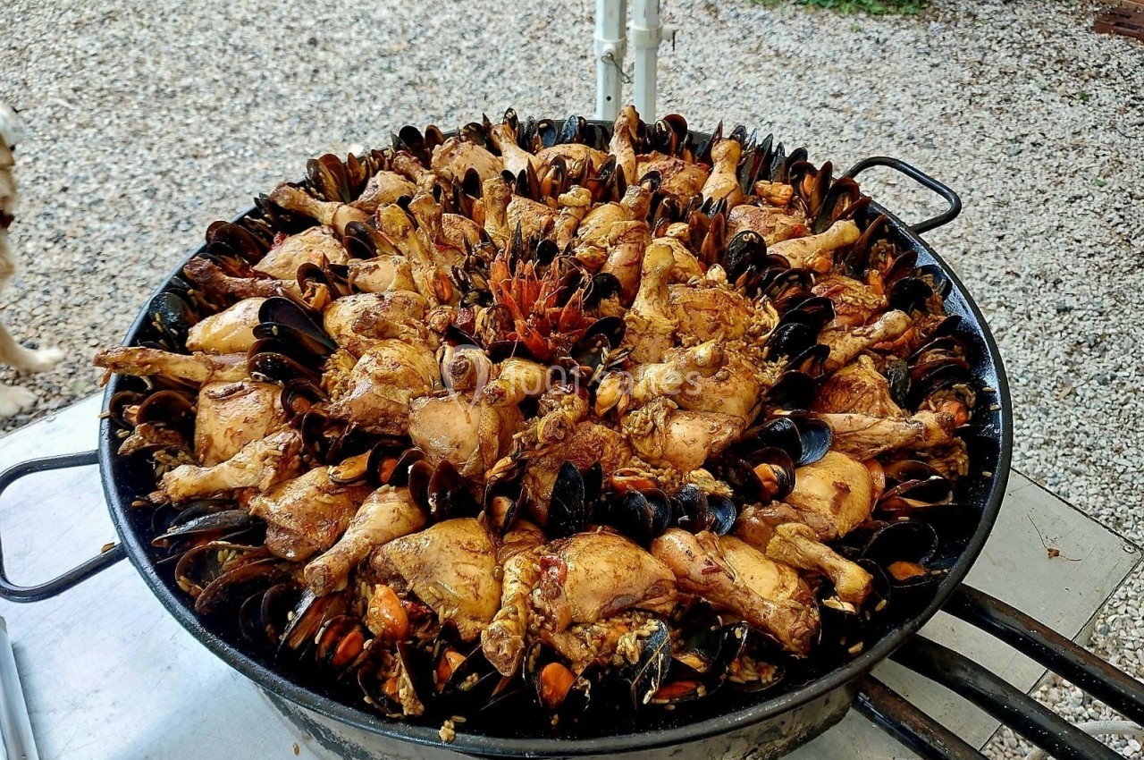 Plat de paella cuisiné avec des morceaux de poulet et des moules, présenté dans une grande poêle en extérieur.