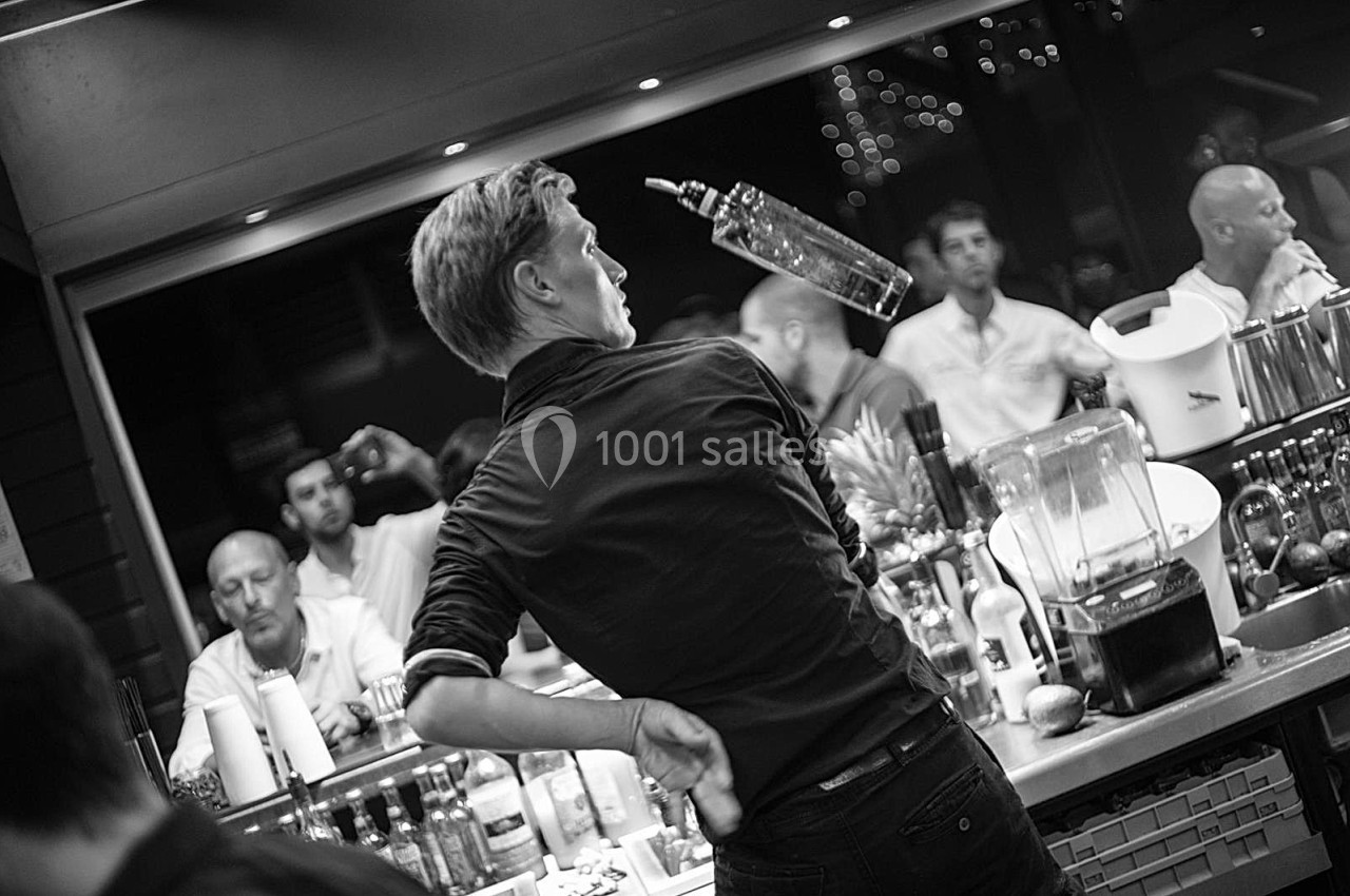 Un barman jongle avec une bouteille dans un bar animé, observé par des clients en arrière-plan.