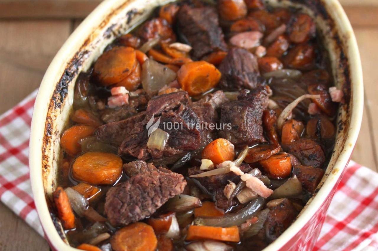 Plat de bœuf mijoté avec des carottes, oignons et lardons dans un plat en céramique sur une nappe à carreaux rouges.