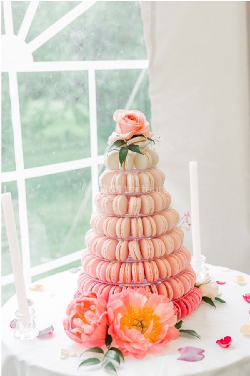 Tour de macarons roses et blancs décorée de fleurs, posée sur une table avec des bougies et des pétales.