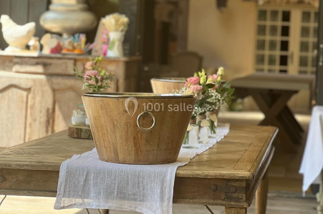 Table en bois décorée avec un seau en bois, des fleurs et une nappe blanche, dans un espace intérieur lumineux.