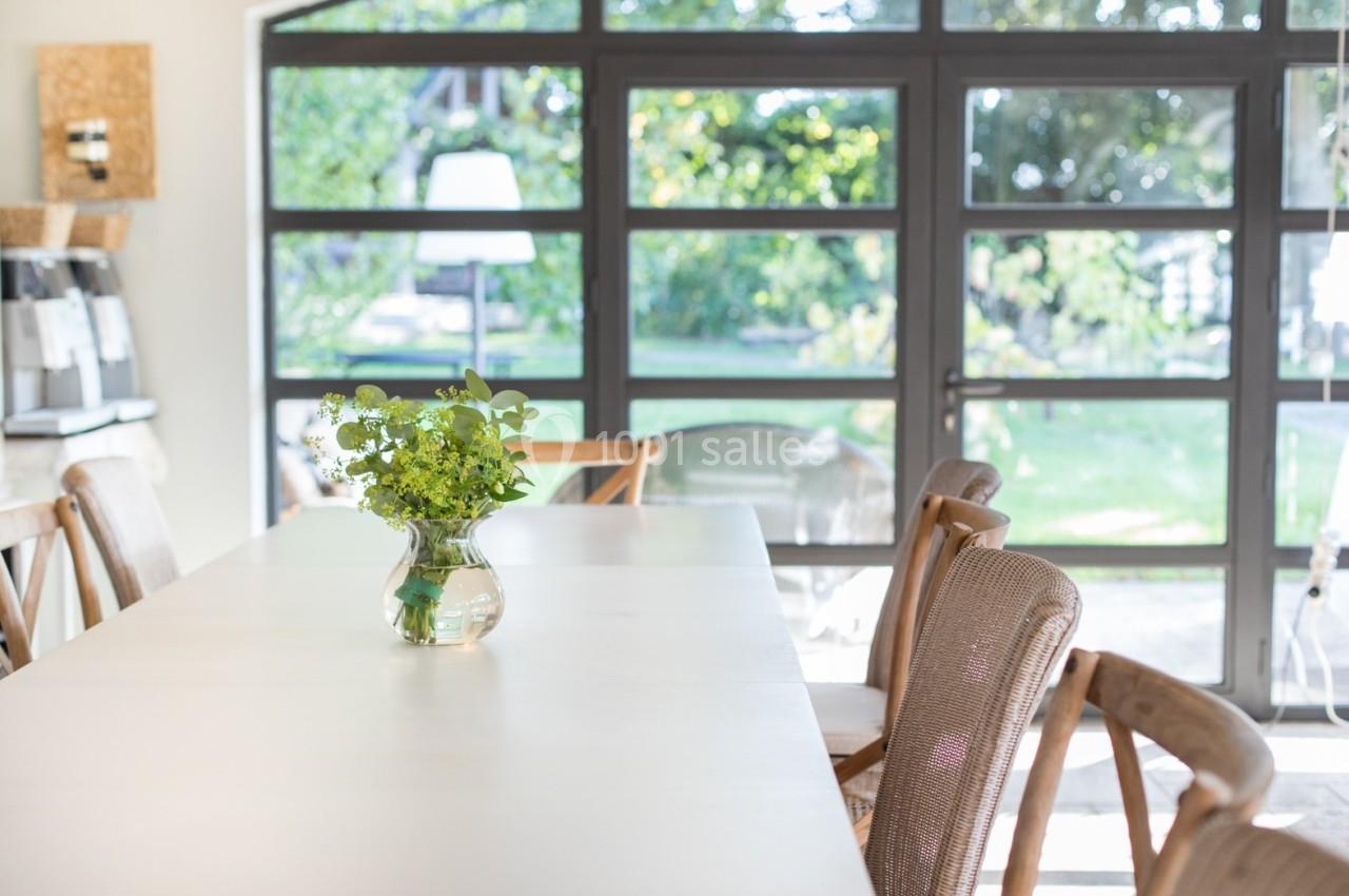 Salle lumineuse avec une grande table blanche, un vase de fleurs vertes et des chaises en bois devant des baies vitrées.