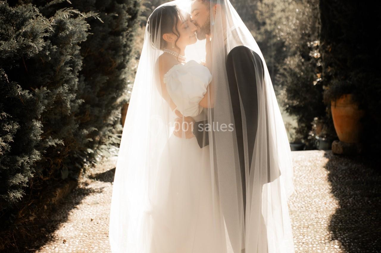 Un couple de mariés s'embrasse sous un voile, debout sur une allée ensoleillée bordée de végétation.