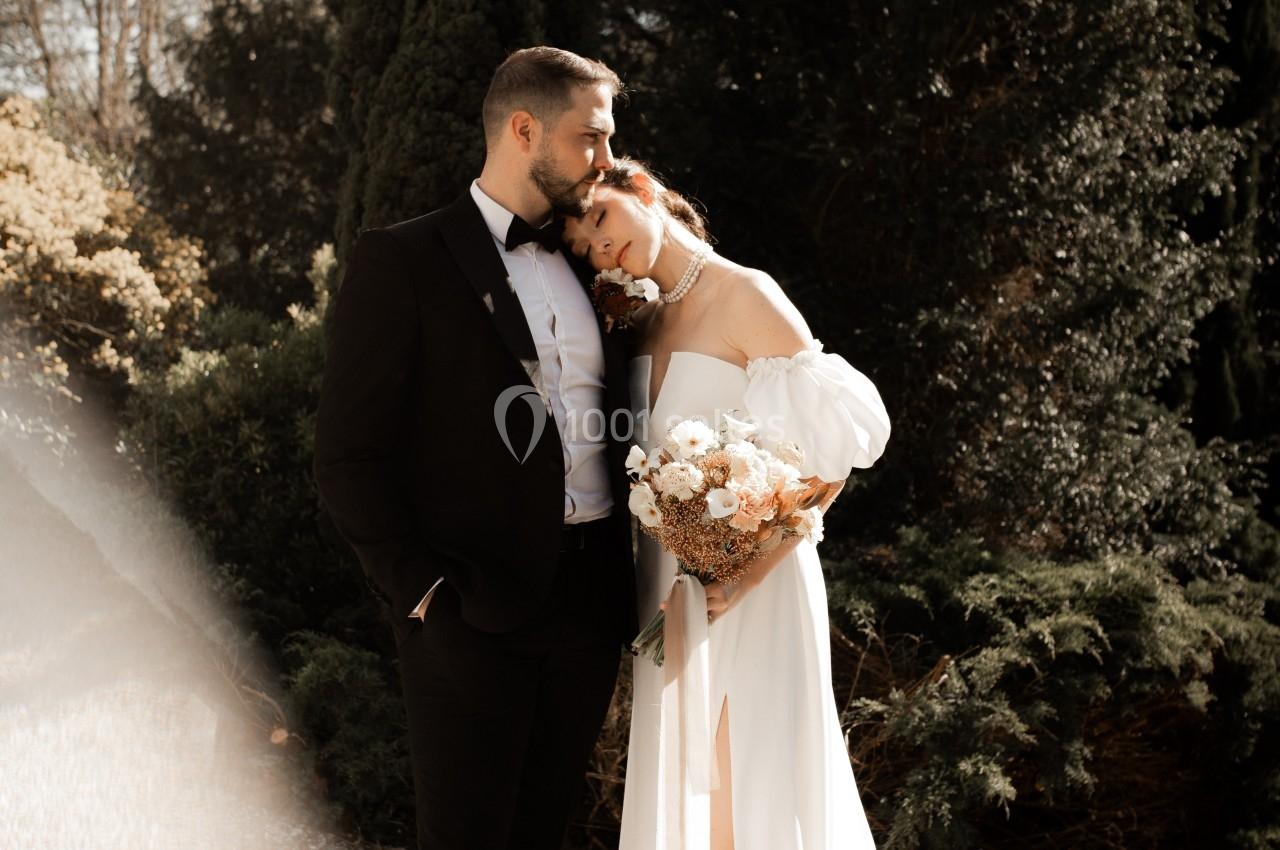 Un couple en tenue de mariage pose dans un jardin ensoleillé, entouré de végétation et d'arbres.