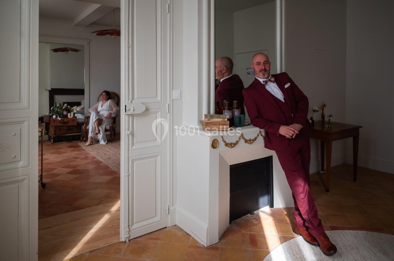 Un homme en costume bordeaux pose près d'une cheminée, une femme assise est visible dans une pièce adjacente.