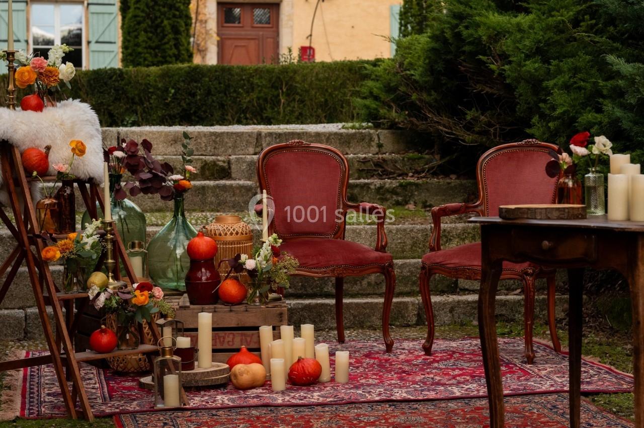 Deux fauteuils rouges et une décoration automnale avec bougies et citrouilles, disposés devant une maison aux volets bleus.