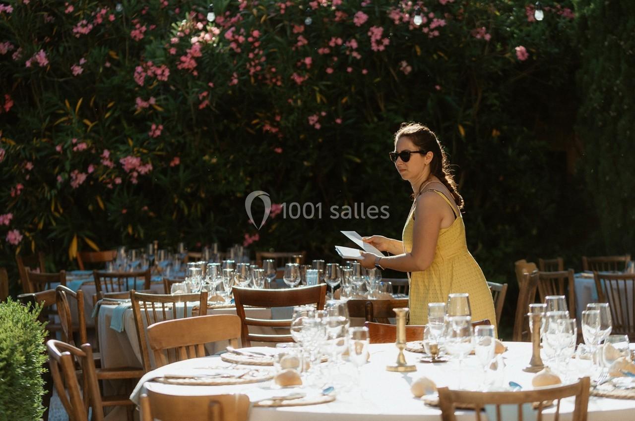 Une femme en robe jaune organise des tables dressées en extérieur, entourées de végétation et de fleurs.