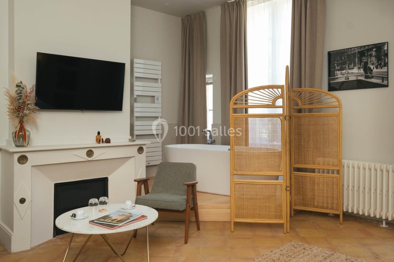 Salon avec cheminée, fauteuil, table basse, paravent en rotin et baignoire près d'une fenêtre avec rideaux beiges.
