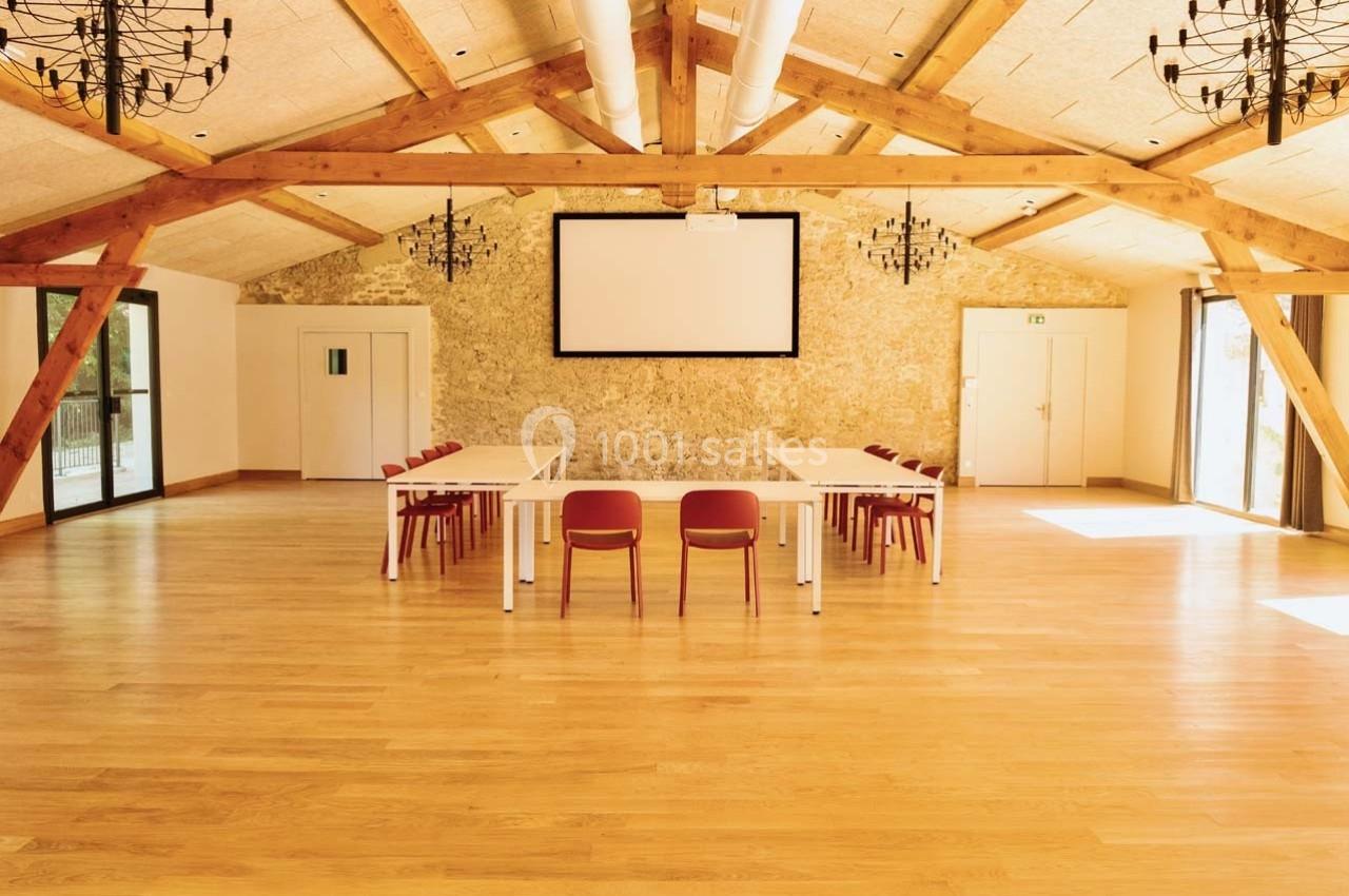 Salle lumineuse avec parquet en bois, tables blanches alignées, chaises rouges et écran de projection au mur.