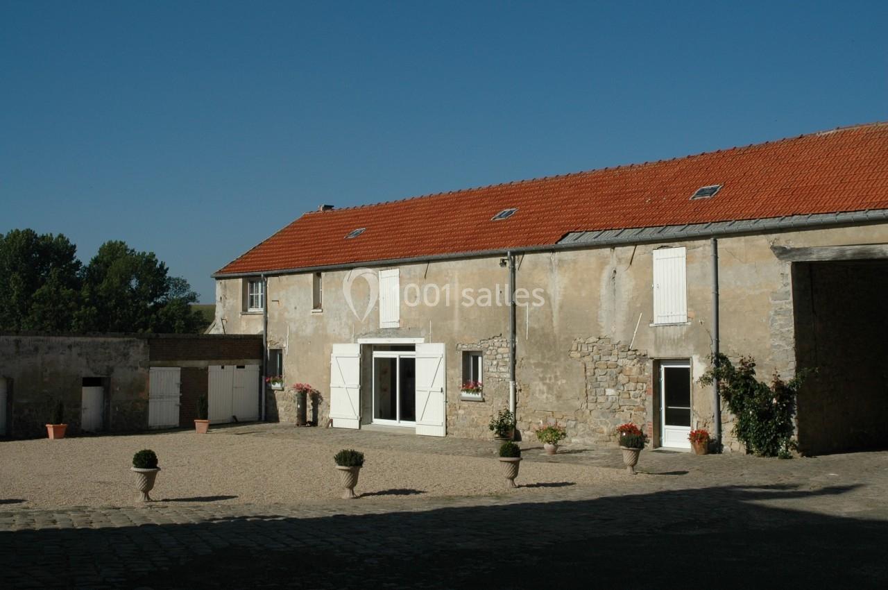 Cour pavée avec un bâtiment en pierre à toit rouge, fenêtres et portes blanches, entourée de pots de fleurs.