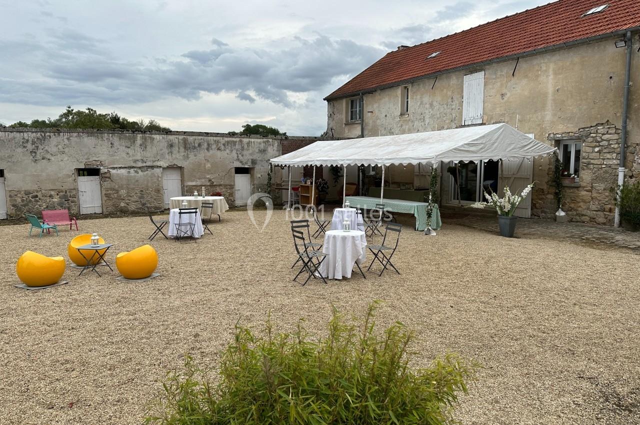 Cour extérieure avec graviers, tables dressées, chaises pliantes, poufs jaunes et une tente blanche adossée à un bâtiment…
