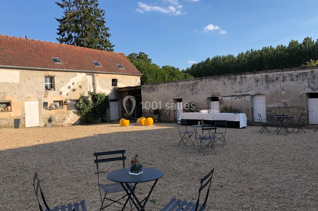 Cour extérieure avec graviers, tables et chaises en métal, bâtiments anciens en pierre et ciel dégagé.