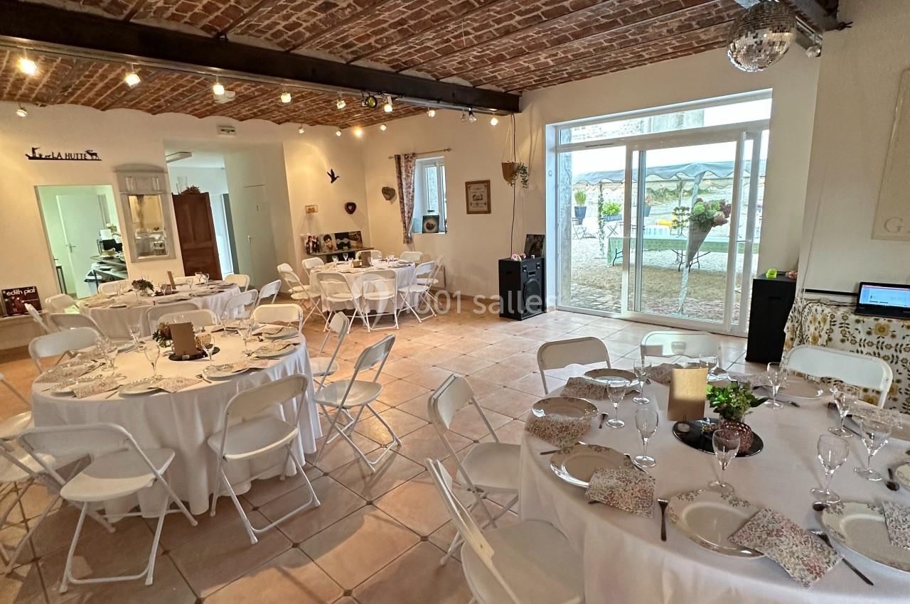 Salle de réception avec tables rondes dressées, chaises blanches pliantes et plafond en briques apparentes.