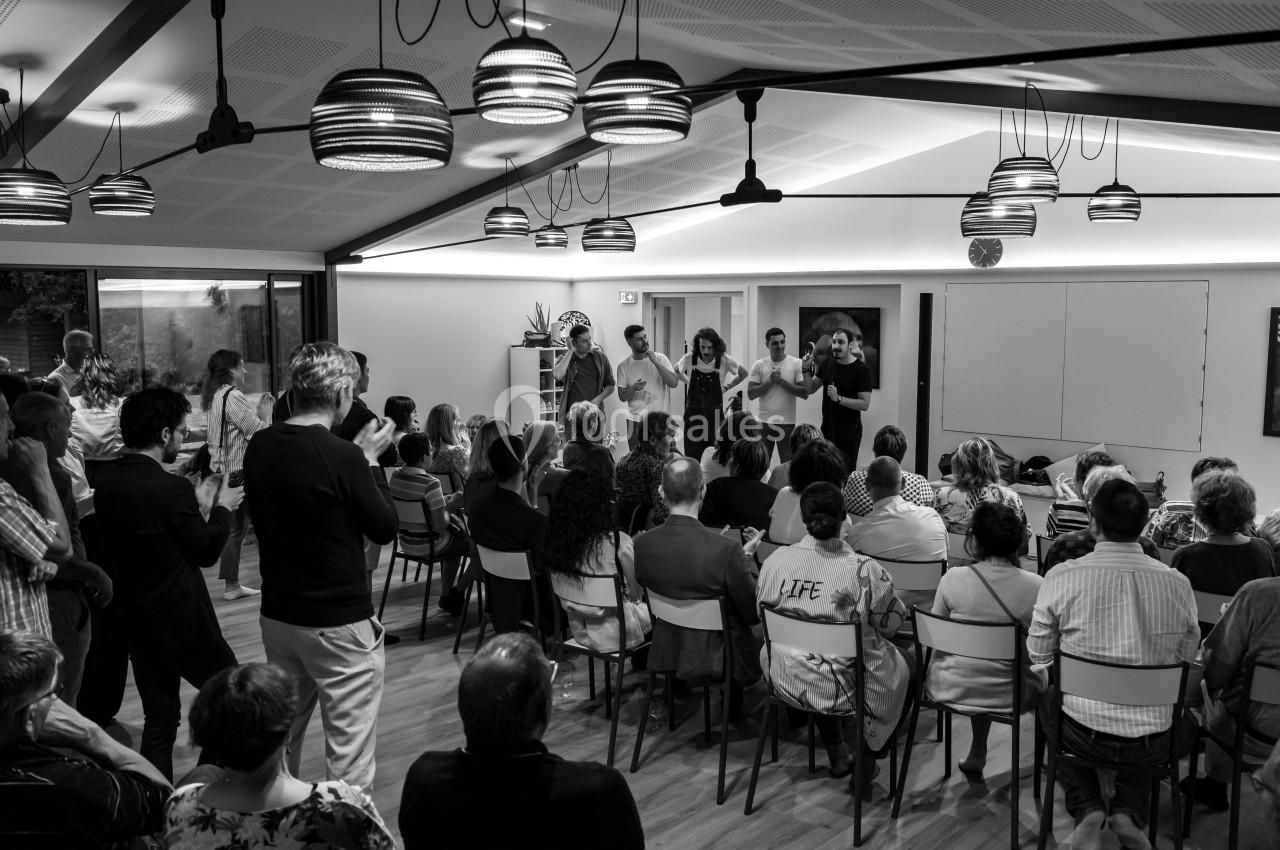 Un groupe de personnes se produit devant un public assis dans une salle lumineuse avec des suspensions au plafond.