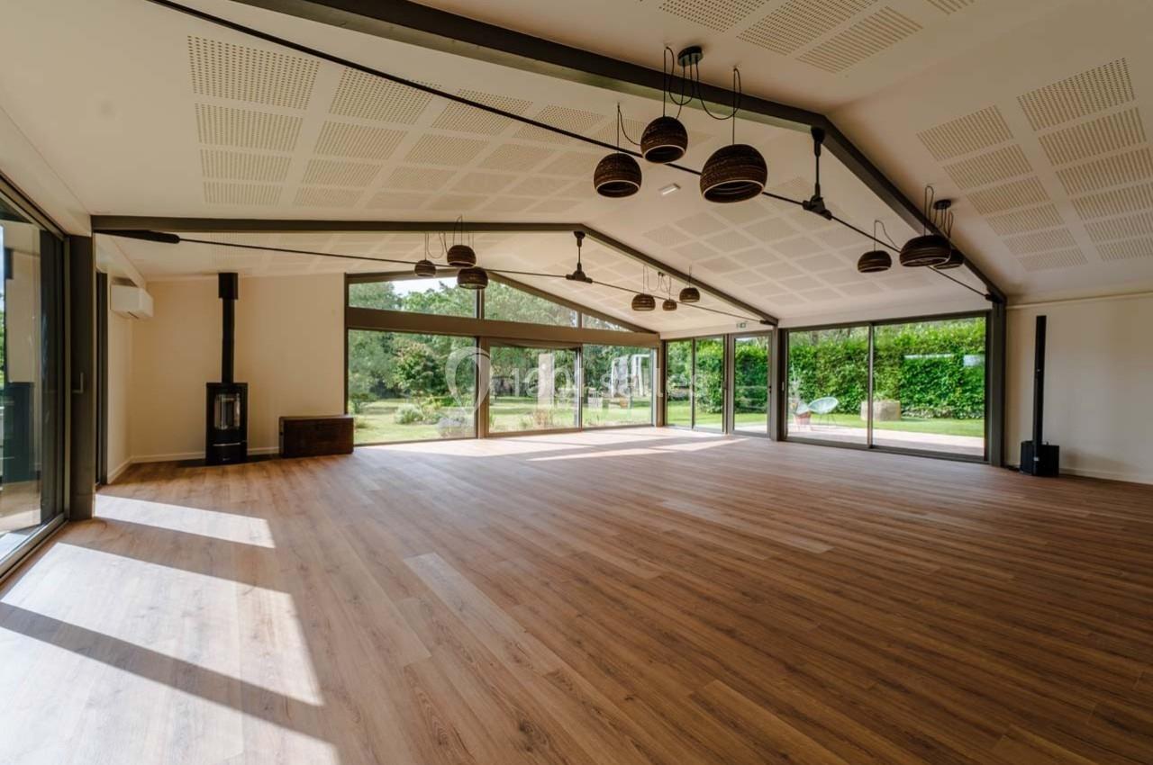 Salle lumineuse avec parquet en bois, grandes baies vitrées donnant sur un jardin verdoyant, et plafond avec luminaires…