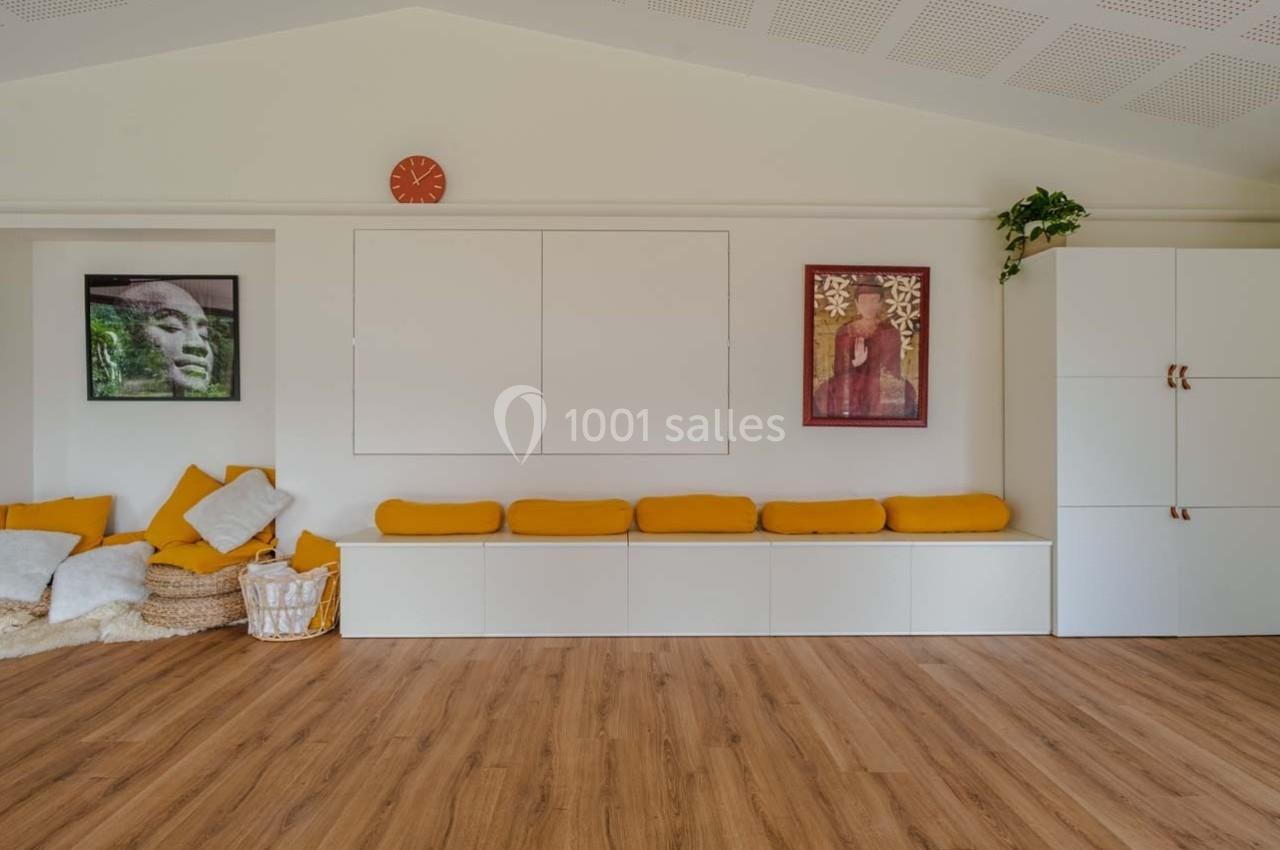 Salle lumineuse avec bancs blancs, coussins jaunes, parquet en bois, décorations murales et plantes.