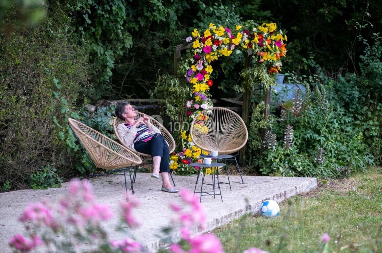Une femme assise sur une chaise en rotin dans un jardin fleuri, près d'une arche décorée de fleurs colorées.